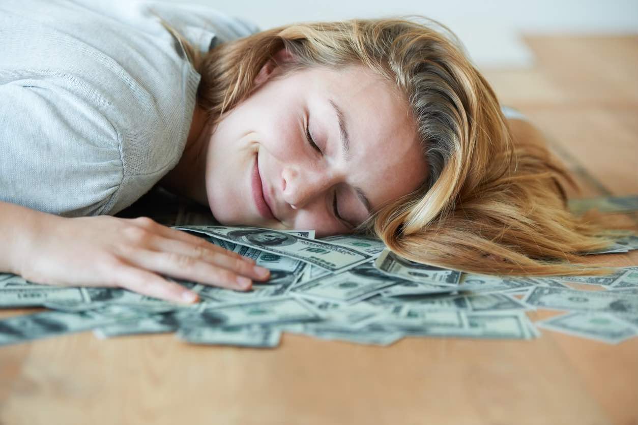 mmm-A smiling girl with her eyes closed lying down on the floor with her head in a pile of money